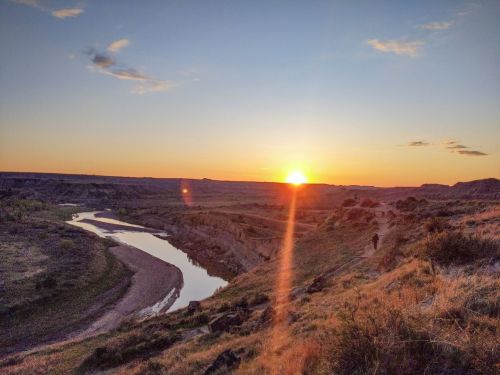 A serene sunset over a winding river, with rolling hills and a lone figure walking along a path.