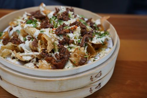 A bamboo steamer filled with crispy nachos topped with seasoned beef, drizzled with sauce and garnished with herbs.
