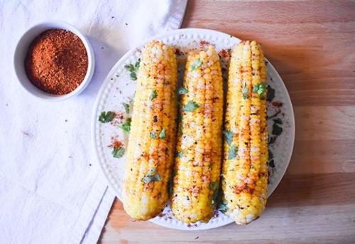 Three grilled corn on the cob topped with herbs and spices, served on a white plate with a small bowl of seasoning.