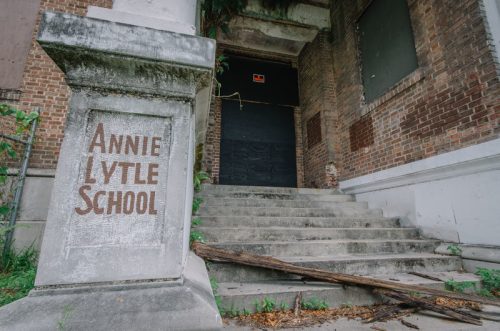 Abandoned building with "Annie Lytle School" sign on a stone pillar and overgrown steps leading to a boarded door.