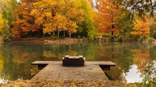 A serene dock extends over a calm lake, surrounded by vibrant autumn trees reflecting in the water.