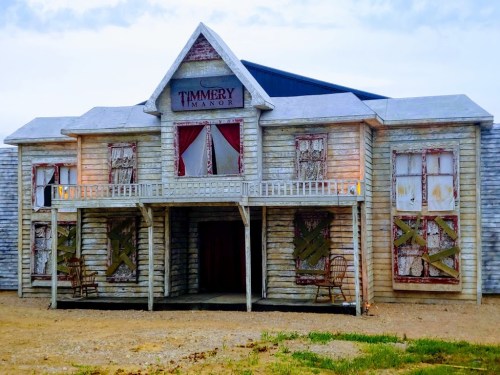 A weathered, two-story building named "Timmery Manor" with boarded windows and a porch, set against a cloudy sky.
