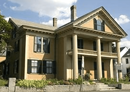A large, historic two-story house with columns, green shutters, and a porch, set against a blue sky.