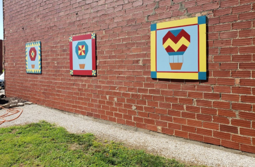 Three colorful quilt block designs mounted on a red brick wall, with a grassy area and gravel path below.