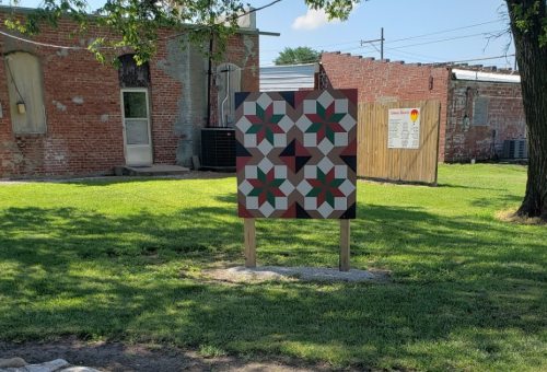 A colorful quilt pattern displayed on a wooden board in a grassy area, with brick buildings in the background.