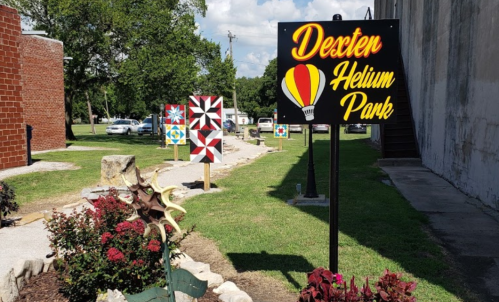 Sign for Dexter Helium Park with colorful quilt squares in the background and flower beds in the foreground.