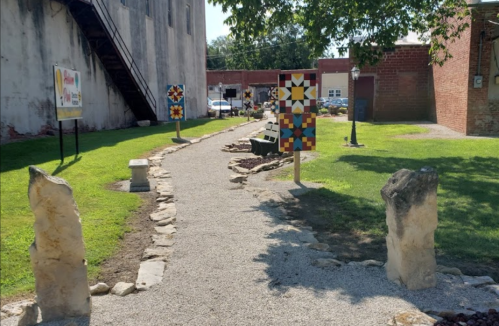 A gravel path lined with colorful quilt block art and stone markers, leading through a green park area.