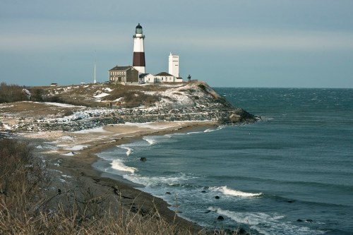 A lighthouse on a rocky shore, with waves crashing against the beach and a cloudy sky overhead.