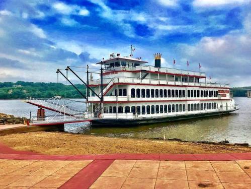 A large riverboat docked at a shore, with a cloudy sky and calm water in the background.