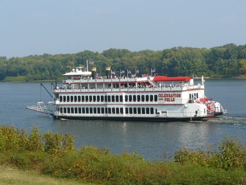 A large white riverboat named "Celebration Belle" sails on a calm river, surrounded by green trees.