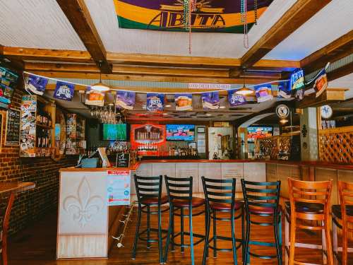 A cozy bar interior with a wooden counter, empty chairs, and colorful decorations, including flags and beer signs.