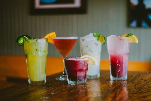 A variety of colorful cocktails on a wooden table, featuring garnishes like lemon and lime.
