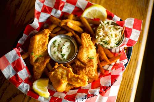 A basket of fried fish with lemon, fries, coleslaw, and dipping sauce, served in a red and white checkered paper.