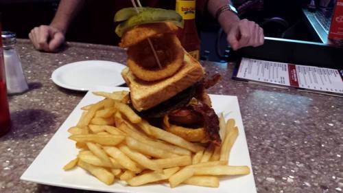 A towering burger with bacon, onion rings, and pickles, served on a plate with a generous side of fries.