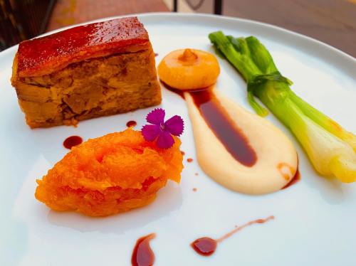 A gourmet plate featuring meatloaf, a creamy sauce, glazed leeks, and a vibrant orange puree, garnished with a flower.