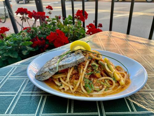 A plate of pasta topped with grilled fish and garnished with a lemon slice, set against a backdrop of red flowers.