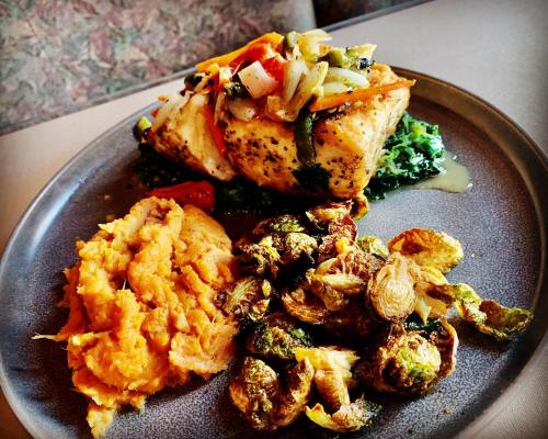 A plate featuring grilled salmon topped with vegetables, served with mashed sweet potatoes and roasted Brussels sprouts.