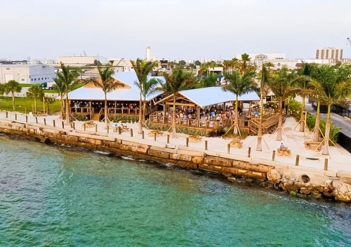 A waterfront restaurant with palm trees, surrounded by water and a walkway, bustling with people enjoying the outdoor space.