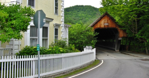 A scenic view of a covered bridge surrounded by greenery and a charming house, with a winding road leading to it.