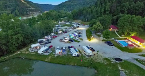 Aerial view of a campground with RVs, cars, and a pool, surrounded by trees and hills at dusk.