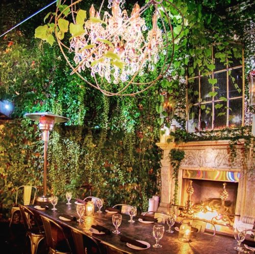 A beautifully decorated outdoor dining area with a chandelier, greenery, and a cozy fireplace.