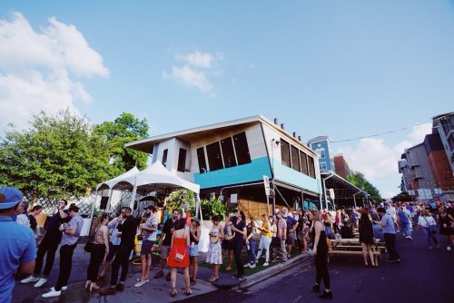 A lively street scene with a crowd gathered outside a colorful building, enjoying an event under a clear blue sky.