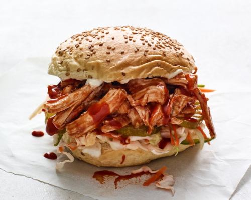 A sesame seed bun filled with shredded BBQ chicken, coleslaw, pickles, and drizzled with sauce.
