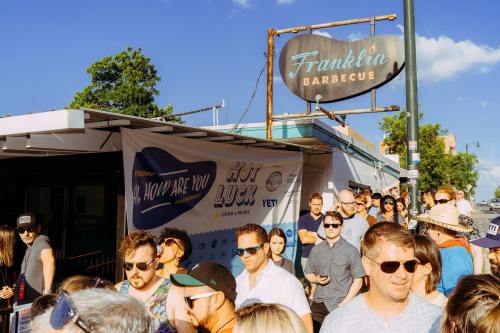 Crowd of people waiting in line outside Franklin Barbecue, with a sunny sky and a promotional banner visible.