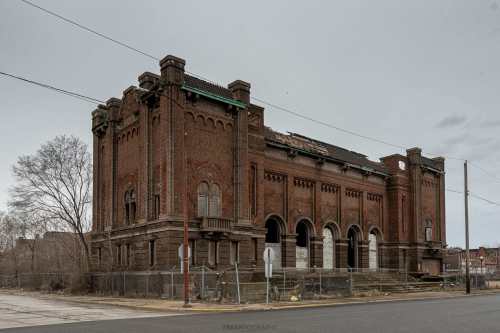 Abandoned brick building with arched windows, overgrown surroundings, and a cloudy sky in the background.