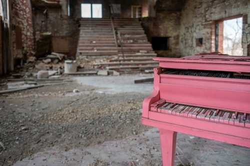 A pink piano sits in a dilapidated room with crumbling walls and a staircase in the background.
