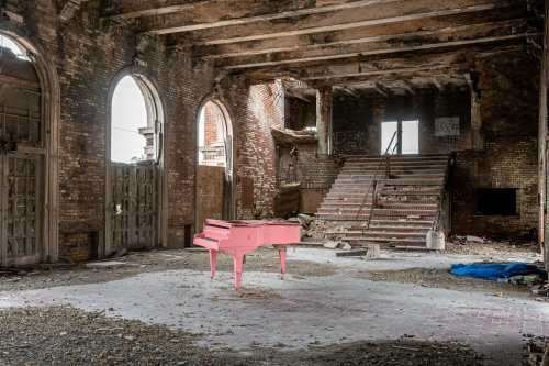 A pink piano sits in the center of a dilapidated, abandoned building with crumbling walls and debris scattered around.