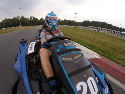 A person driving a go-kart on a racetrack, wearing a helmet and racing gear, with a blurred background of the track.