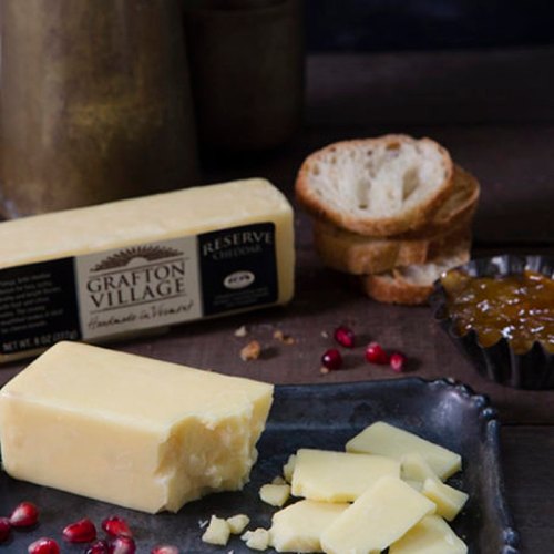 A block of Grafton Village cheese with slices, crackers, and a small dish of jam on a rustic wooden surface.