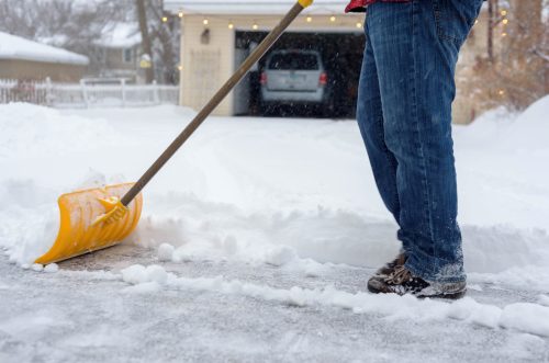 A person shoveling snow from a driveway, wearing jeans and boots, with a yellow shovel in hand. Snow is falling.