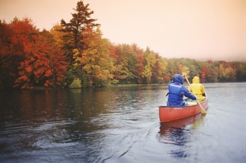 Two people in colorful raincoats paddle a canoe on a calm lake surrounded by vibrant autumn foliage.