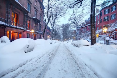 A snowy street lined with brownstone buildings, street lamps glowing, and cars buried under snow.