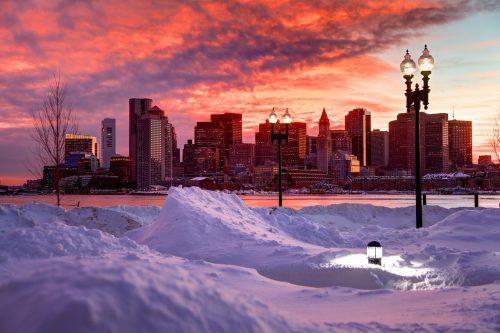 A snowy landscape at sunset, with a city skyline in the background and street lamps illuminating the scene.