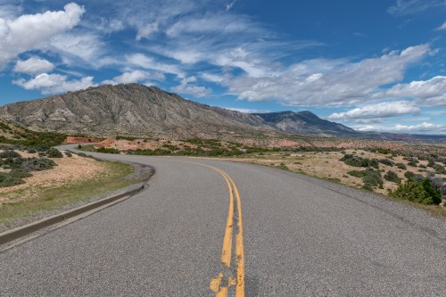 A winding road through a scenic landscape with mountains and blue skies, featuring fluffy clouds.