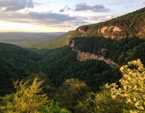 A scenic view of a lush green valley surrounded by mountains under a cloudy sky during sunset.