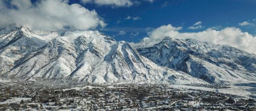 Snow-covered mountains rise above a valley, with a small town nestled at the base under a partly cloudy sky.