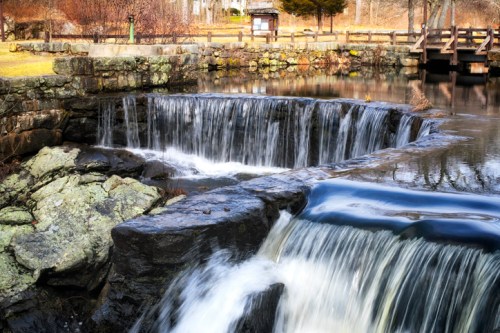 A serene waterfall cascades over rocks into a calm river, surrounded by trees and a stone bridge in a peaceful park setting.