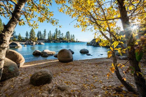 A serene lakeside scene with golden autumn leaves, smooth boulders, and calm water under a clear blue sky.