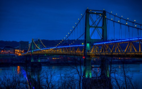 A brightly lit bridge spans a river at night, with colorful lights reflecting on the water and a city skyline in the background.