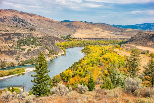 A winding river flows through a valley surrounded by autumn-colored trees and rolling hills under a cloudy sky.