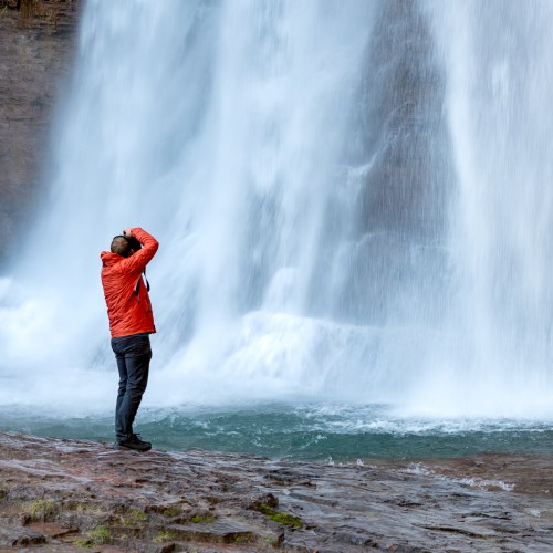 A person in a red jacket stands by a waterfall, capturing the scene with a camera amidst the mist.