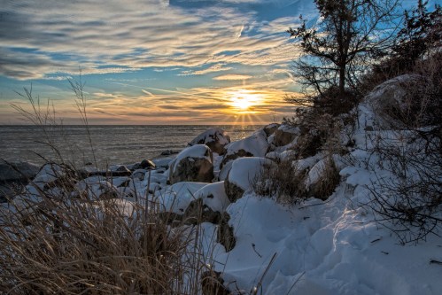 A serene winter landscape featuring snow-covered rocks and grass, with a sunset over a frozen lake and cloudy sky.