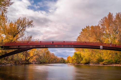 A red bridge spans a calm river, surrounded by vibrant autumn trees under a cloudy sky.