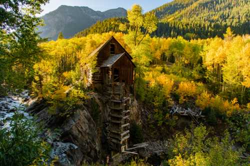 A rustic wooden cabin on a rocky ledge, surrounded by vibrant autumn foliage and mountains in the background.