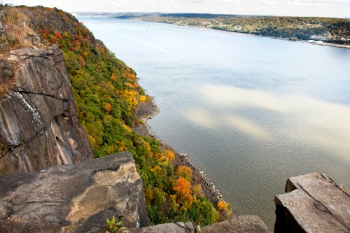 A scenic view of a river bordered by colorful autumn foliage and rocky cliffs under a partly cloudy sky.