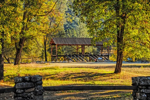 A rustic pavilion surrounded by trees in a serene outdoor setting, with a grassy area and stone features in the foreground.